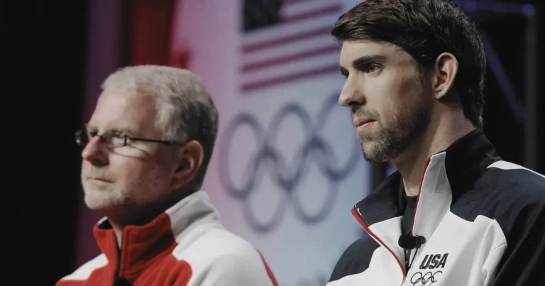 Michael Phelps and coach Bob Bowman at a press conference, both wearing USA Olympic team jackets, with focused expressions.