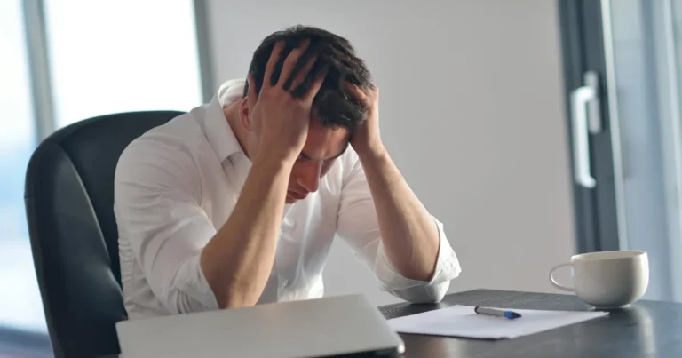 Frustrated CFA candidate sitting at a desk, holding his head in his hands while preparing for exams, with a laptop, pen, and coffee cup on the table.